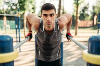 Athletic man in headphones doing exercise on parallel bars, front view, outdoor fitness workout. Strong sportsman on sport training in park