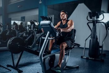 Muscular athlete training legs on exercise machine. Active sport exercises in gym. Muscular athlete training legs on exercise machine