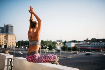 Young woman sitting in yoga pose, city on background. Yogi training outdoors, relaxation exercise