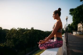 Young woman sitting in yoga pose, city on background. Yogi training outdoors, relaxation exercise. Young woman in yoga pose, city on background