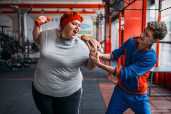 Trainer takes hotdog at fat woman, motivation, hard workout in gym. Calories burning, obese female person in fitness club, fat-burning, sport against unhealthy food