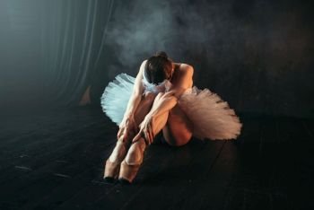 Female ballet performer in white dress sits on the floor, body flexibility. Ballerina training in dancing class