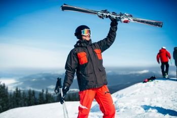 Skier in helmet and glasses holds skis and poles in hands, blue sky and snowy mountains on background. Winter active sport, extreme lifestyle. Downhill skiing