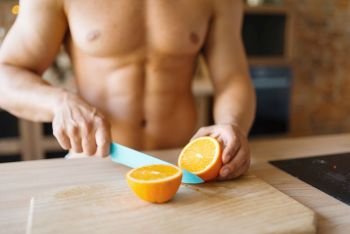 Man with naked body cuts orange on the kitchen. Nude male person preparing breakfast at home, food preparation without clothes