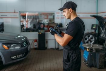Auto mechanic in uniform holds inspection report, tire service. Worker repairs car tyre in garage, professional automobile inspection in workshop, vehicle on lift jack on background. Mechanic holds inspection report, tire service