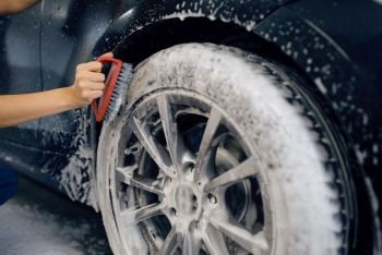 Female washer with brush in hand cleans wheel in foam, car wash. Woman washes vehicle, carwash station, car wash business. Female washer with brush cleans wheel, car wash
