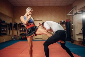 Female person makes a kick in the groin, self defense workout with male personal trainer, gym interior on background. Woman on training, self-defense practice