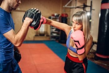 Female kickboxer on workout with male personal trainer in pads, gym interior on background. Woman boxer makes hand punch on training, kickboxing practice