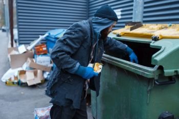 Male bearded beggar searching food in trashcan on city street. Poverty is a social problem, homelessness and loneliness, alcoholism and drunk addiction. Male bearded beggar searching food in trashcan