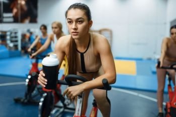 Woman drinks water on a stationary bike in gym. People on fitness workout in sport club, athletic girls in sportswear on training indoors. Woman drinks water on a stationary bike in gym