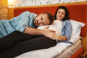 Husband listens to baby in pregnant wife’s stomach at home, bedroom interior on background. Pregnancy, prenatal period. Expectant mom and dad are resting on sofa, health care. Husband listens to baby in pregnant wife’s stomach