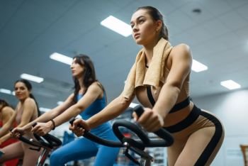 Group of sportive women doing exercise on stationary bikes in gym. People on fitness workout in sport club, athletic girls in sportswear on training indoors. Sportive women on stationary bikes in gym