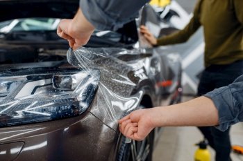 Male worker applies car protection film on front fender. Installation of coating that protects the paint of automobile from scratches. New vehicle in garage, tuning procedure. Worker applies car protection film on front fender
