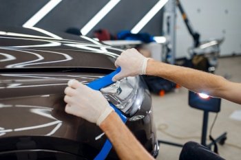 Male worker prepares car surface to applying of protection film. Installation of coating that protects the paint of automobile from scratches. Vehicle in garage, detailing. Worker prepares car to applying of protection film
