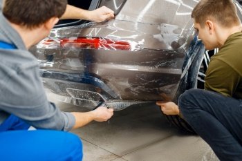 Two workers applies car protection film on rear bumper. Installation of coating that protects the paint of automobile from scratches. New vehicle in garage, tuning procedure. Two workers applies car protection film on bumper