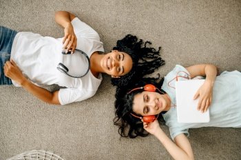 Two women in headphones enjoys listening to music lying on the floor. Pretty girlfriends in earphones relax in the room, sound lovers resting on couch. Two women in headphones enjoys listening to music
