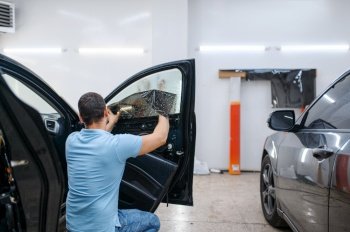 Male specialist installs wetted car tinting, tuning service. Mechanic applying vinyl tint on vehicle window in the garage. Specialist installs wetted car tinting, tuning