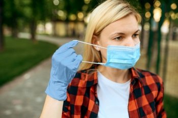 Young woman in gloves puts mask on her face in park, quarantine. Female person walking during the epidemic, health care and protection, pandemic lifestyle. Woman in gloves puts mask on her face, quarantine