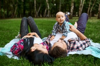 Mother, father and little baby leisures on grass. Mom and dad with male kid on lawn, parents on picnic with child in the forest, family happiness. Mother, father and little baby leisures on grass
