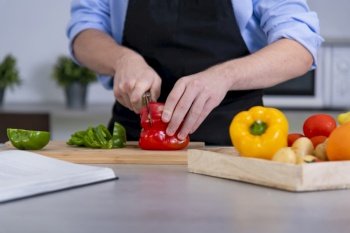 Close up of a young male cutting a red bell pepper on a cutting board surrounded by assorted vegetables on an out of focus background. Safety and cooking at home concept.. a young male cutting a red bell pepper on a cutting board surrounded by assorted vegetables