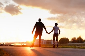 Defocused couple riding on roller skates, holding hands on the street during sunset in summer, back view from the bottom. Copy space. Active lifestyle concept. 