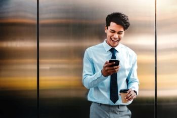 Portrait of a Happy Young Businessman Using Mobile Phone in the Urban City. Lifestyle of Modern People. Front View. Standing by the Wall with Coffee Cup