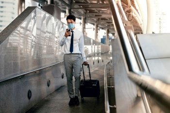 Young Asian Businessman Wearing a Surgical Mask and Using a Smart Phone while Walking with Suitcase in the Airport or Public Transportation Station.Healthcare in New Normal Lifestyle Concept