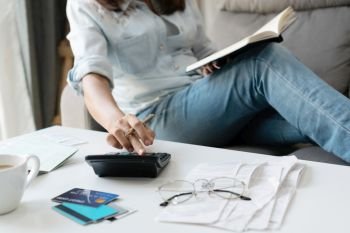 Pretty young Asian woman using calculator while holding notebook to note home expenses and taxes in living room at home. 