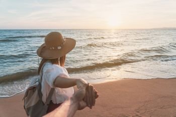 Young woman traveler holding man’s hand and looking beautiful sunset on the beach, Couple vacation travel summer concept