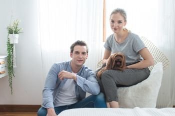 Young happy caucasian couple with smile sit on sofa  hug dog on knee in bedroom at home,indoors lifestyle concept.