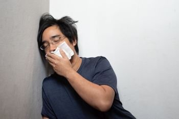 Male Asian young man wearing surgical mask feeling sick headache and coughing leaning on wall isolated on white background.Wuhan coronavirus (COVID-19) outbreak prevention. Health care concept