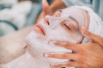 Cosmetologist applying rejuvenating facial mask onto woman’s face in beauty salon. . Rejuvenating Facial Skin Mask Treatment