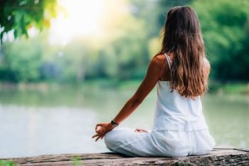 Woman doing yoga by the lake, sitting in lotus pose with fingers in mudra position.