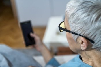 Using Blue-Light Blocking Glasses in the Evening for Better Sleep. Mature Woman lying in bed, holding smart phone . Mature Woman Using Blue-Light Blocking Glasses in the Evening to Minimize Sleep Disruption