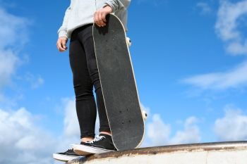 Close up of unrecognizable young man holding skateboard in the park on blue sky background .. Close up of unrecognizable young man holding skateboard in the park on blue sky background.