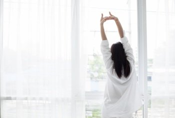Asian woman waking up in her bed fully rested and open the curtains in the morning to get fresh air.