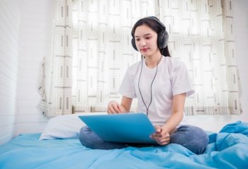 Asian happy young woman wear headphones and listening music song and watching website for shopping online from laptop  on bed at home