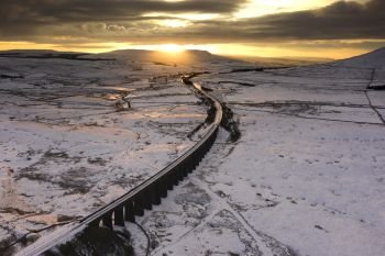 Winter Ariel picture of Yorkshire landmark Ribblehead Viaduct, North Yorkshire, The Ribblehead Viaduct or Batty Moss Viaduct carries the Settle–Carlisle railway across Batty Moss in the Ribble Valley at Ribblehead, in North Yorkshire, England. The viaduct, built by the Midland Railway, is 28 miles (45 km) north-west of Skipton and 26 miles (42 km) south-east of Kendal. It is a Grade II* listed structure.[1] Ribblehead Viaduct is the longest and the third tallest structure on the Settle–Carlisle line.



The viaduct was designed by John Sydney Crossley, chief engineer of the Midland Railway, who was responsible for the design and construction of all major structures along the line. The viaduct was necessitated by the challenging terrain of the route. Construction began in late 1869. It necessitated a large workforce, up to 2,300 men, most of whom lived in shanty towns set up near its base. Over 100 men lost their lives during its construction. The Settle to Carlisle line was the last main railway in Britain to be constructed primarily with manual labour.