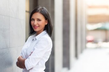 Attractive Hispanic female with arms crossed and looking at camera over building background at outdoor. Smiling Beautiful woman in white shirt standing with confident expression and copy space. 