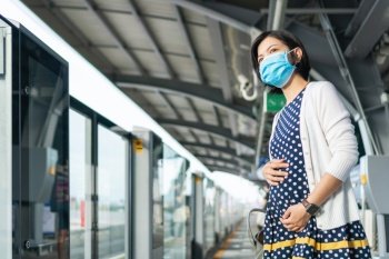 Asian Pregnant Woman in face mask waiting commuter skytrain for traveling to work. Young mother life in public transportation during pandemic virus. Health care in New normal and social distancing.