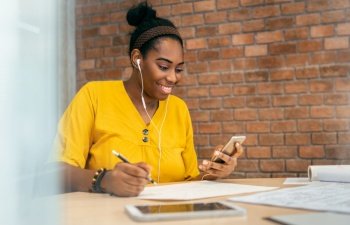 African woman designer using smartphone and earphones for listening to music while working at workplace. African-American Creative Female in Yellow shirt enjoying online video chat with mobile phone