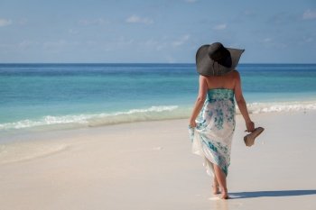 Maldives, young woman walking along the beach with sunhat and high heels on the hand