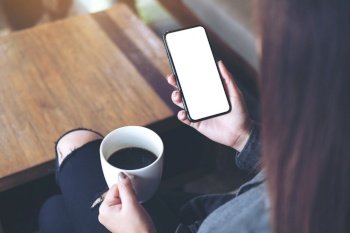 Mockup image of a woman sitting cross legged , holding black mobile phone with blank white desktop screen on thigh while drinking coffee in cafe