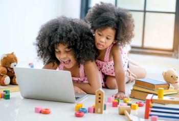 Two young African girls play with notebook computer among toys, doll and book in front of glass window.