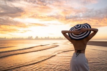 Woman in top bikini and white long pant wearing hat on the beach with a beautiful sunrise or sunset in background.