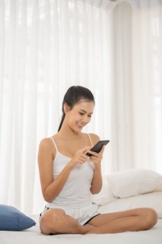 Young Woman Using Mobile Phone While Sitting On Bed At Home