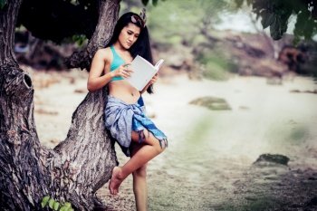 Young Woman Reading Book While Leaning On Tree At Beach