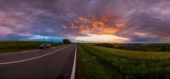 Spring rapeseed yellow fields, cloudy sunset evening sky, rural hills and regional road. Natural seasonal, travel, weather, countryside beauty concept scene. Car unrecognizable.