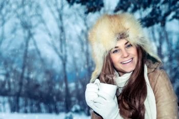 Beautiful happy smiling woman with cup of coffee standing over beautiful winter park background, wearing warm hat and having a hot drink, enjoying wintertime vacation