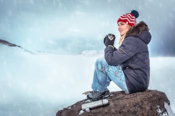 Traveler woman sitting on the big stone with mug of hot tasty mulled wine in hands enjoying view on snowy mountains, having fun outdoors in cold wintertime day, active winter vacation